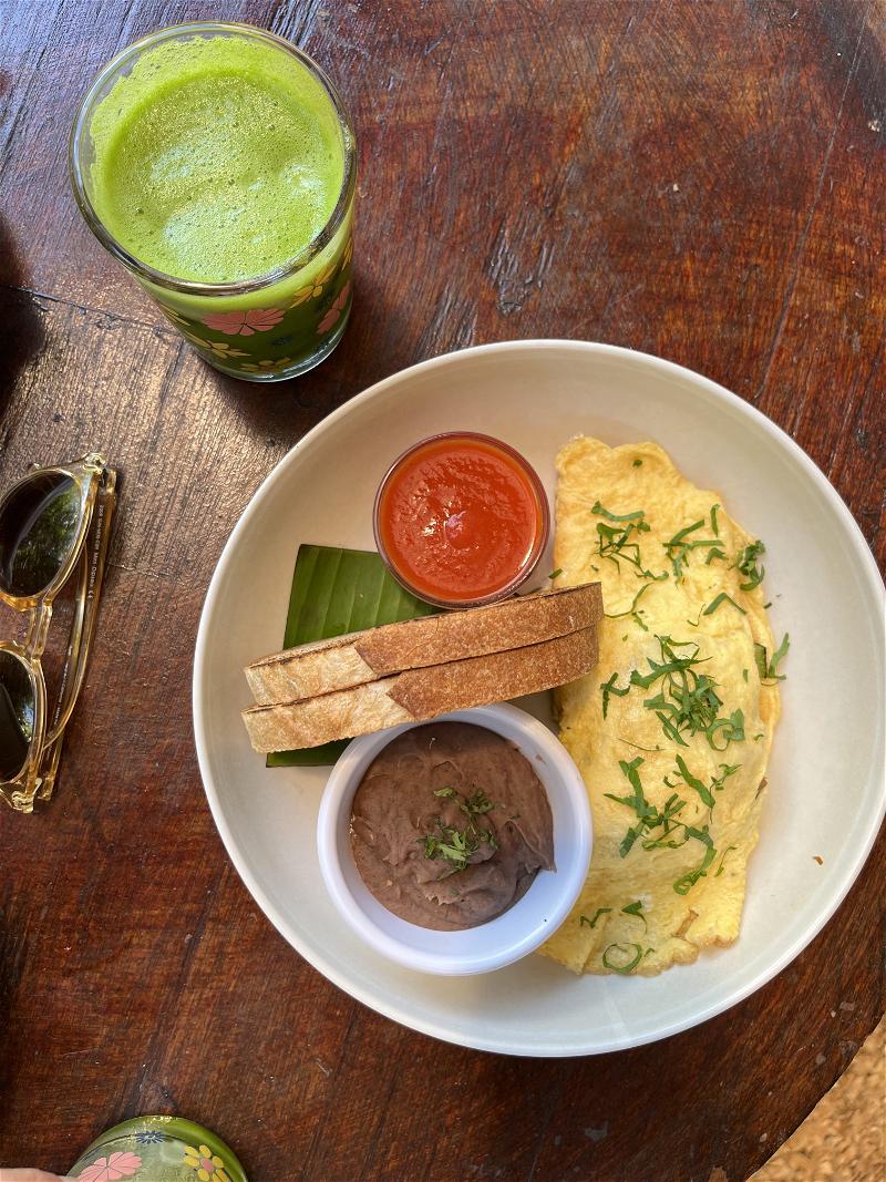 Two slices of bread, an egg omelet and refried black beans on a white plate on a wooden table with a green juice.