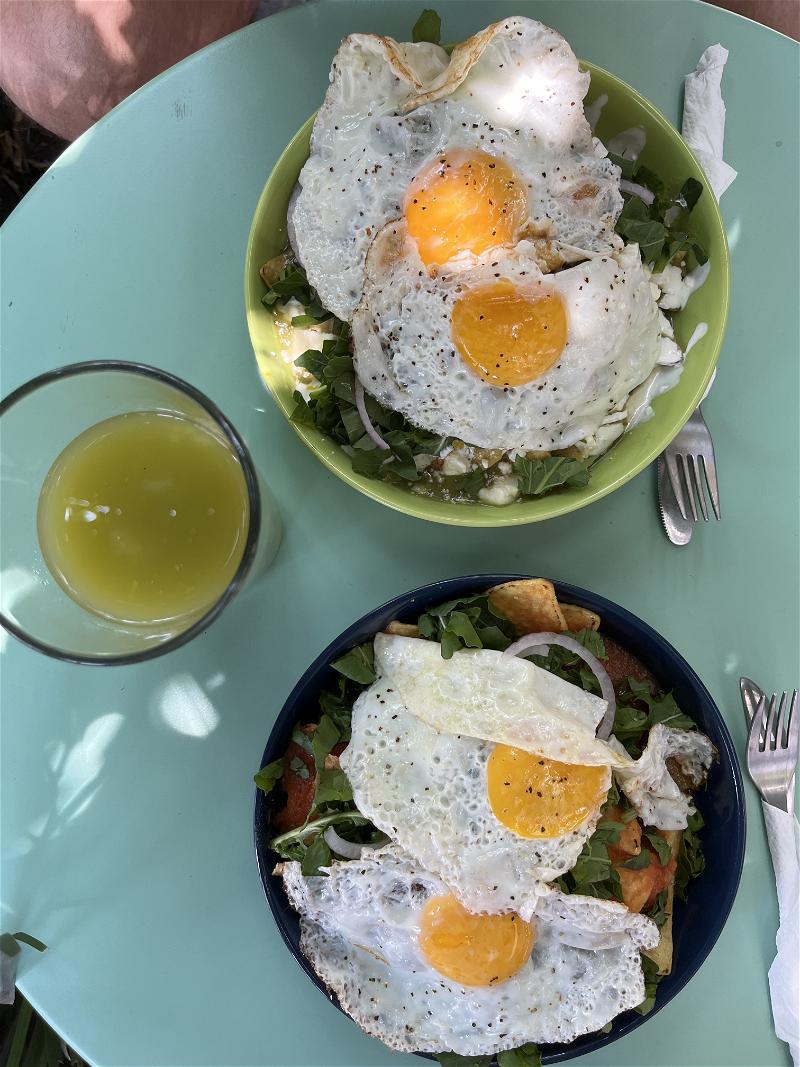 Two plates of chilaquiles with two fried eggs and a green juice on a blue table in Mexico City.