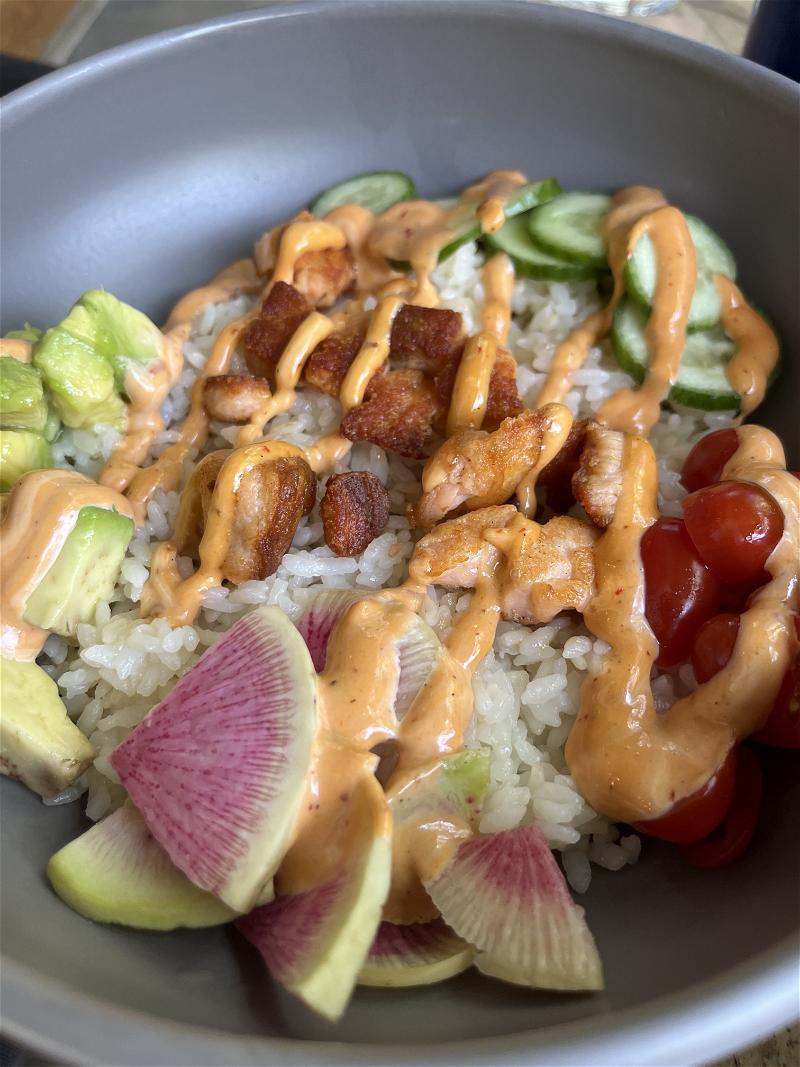 Bowl of poke containing white rice, cucumbers, raddish, cherry tomatoes, salmon and avocado with spicy mayo drizzled on top.
