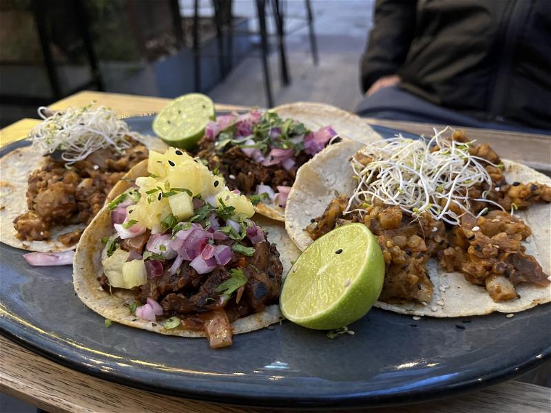 Four vegan tacos on corn tortillas and two sliced limes on a blue plate and a wooden table in Mexico City.