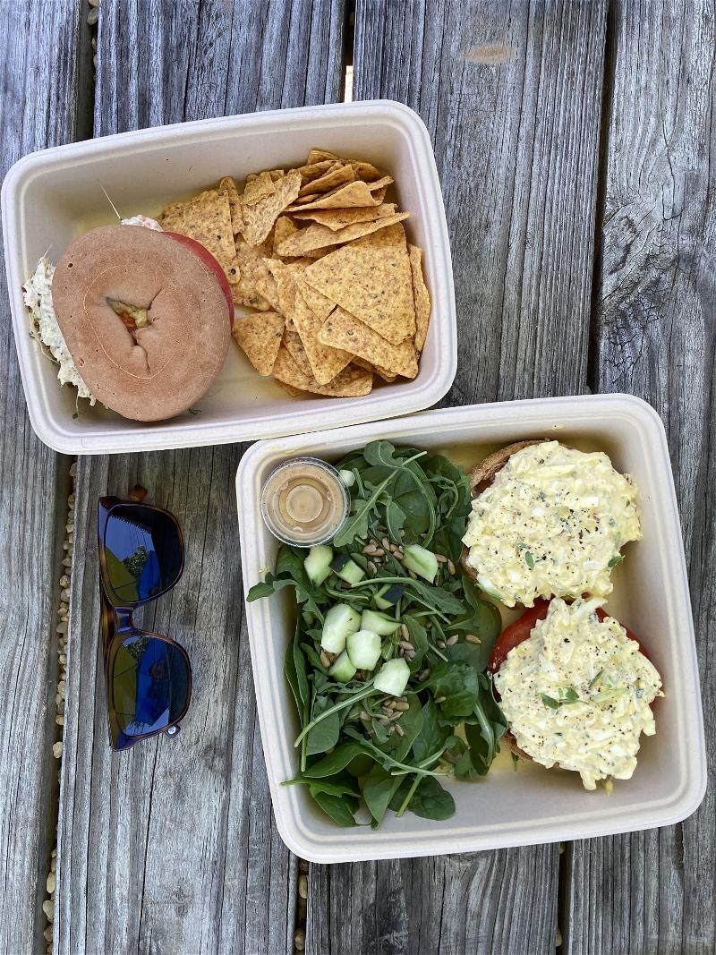Two carry-out lunches of bagels, chips, and egg salad on top of a wooden picnic table with a pair of sunglasses.