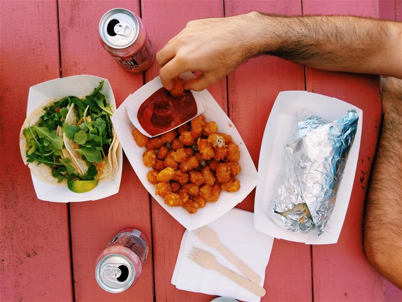A hand dipping tater tots in ketchup in a white disposable tray, with two tacos and a wrapped burrito on a pink picnic table.