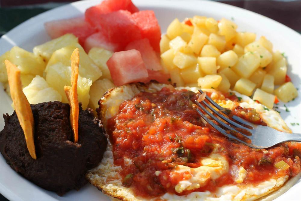 Typical Guatemalan breakfast of black beans, a fried egg on a tortilla, fried potatoes and watermelon with pineapple on a white plate.