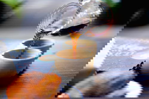 Pouring a stream of black coffee from a glass carafe on a wooden table into a small white cup
