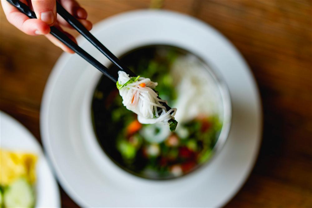 White vermicelli noodles looped on black chopsticks being lifted over a silver bowl on a white plate