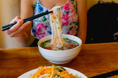 Woman wearing a floral tank top using black chopsticks to lift flat rice pho noodles out of a pho soup