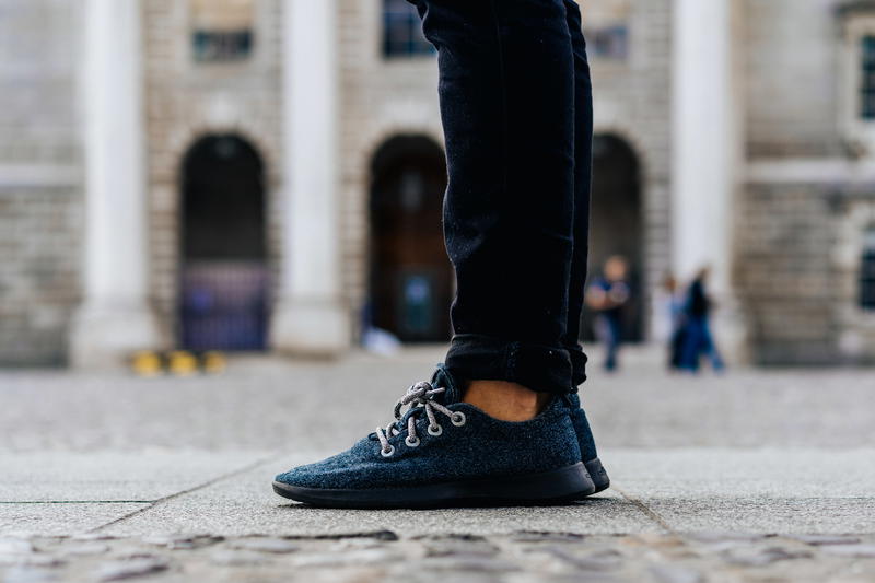 Man wearing black jeans and dark gray Allbirds on a stone sidewalk