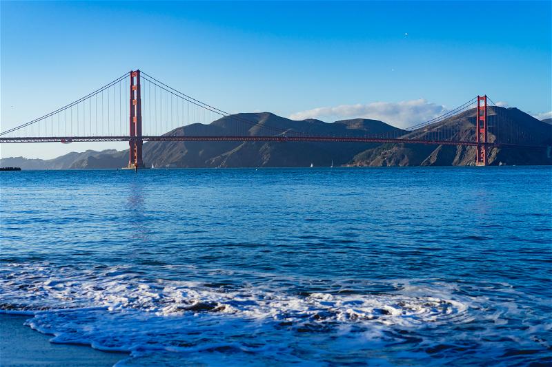 The Golden Gate Bridge on a clear day with mountains in the background and seafoam in the foreground