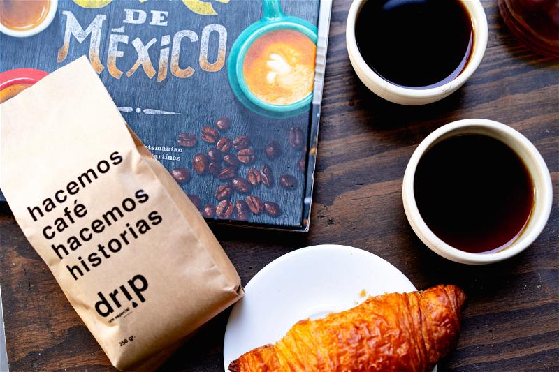 Two small white cups of black coffee, a croissant on a white plate and a brown paper bag of craft coffee in Mexico City Drip Cafe