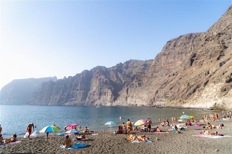 Playa los Gigantes scene with tourists on a black sand volcanic beach and massive brown cliffs overlooking the sea