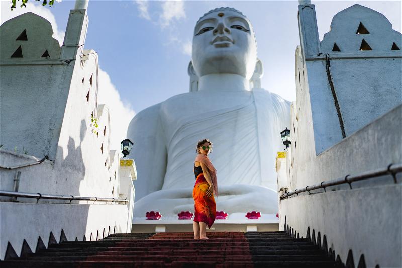A woman wearing a red and orange sarong walks up red steps at the big white buddha of Kandy Sri Lanka