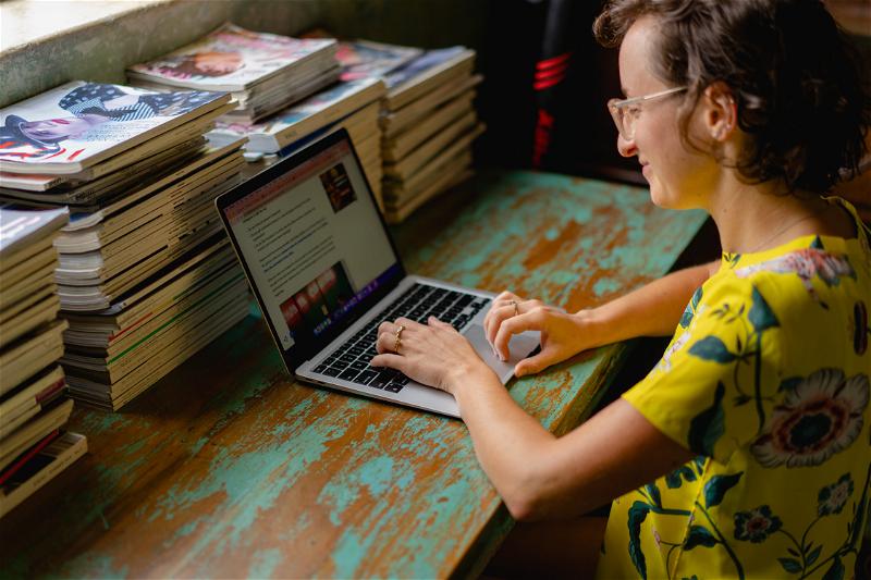 A woman working on a laptop in front of a pile of books.