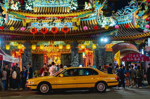 Red lanterns with Chinese symbols hanging in front of a lit-up Buddhist temple with a yellow taxi out front