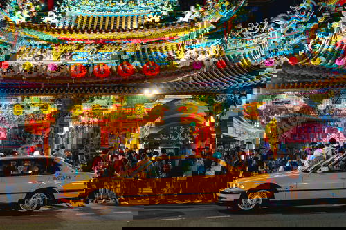 Red lanterns with Chinese symbols hanging in front of a lit-up Buddhist temple with a yellow taxi out front