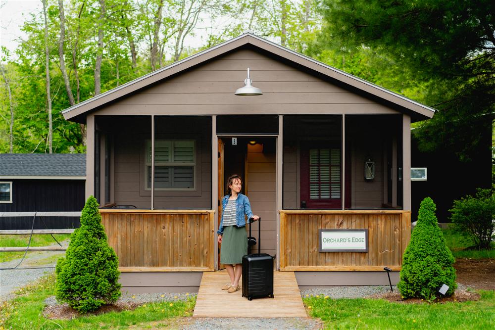 A woman in front of a small house in The Catskills with a suitcase.
