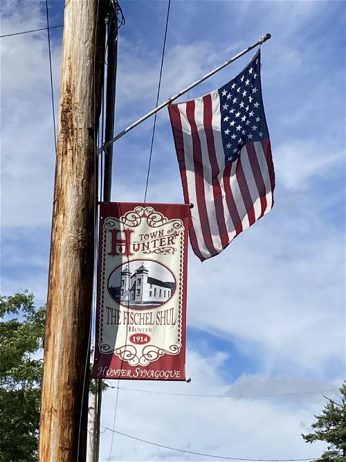An American flag posted on a wooden pole with a flag that reads Town of Hunter and the Fischel Shul, 1914