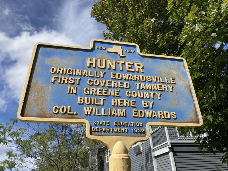 A blue sky with clouds and green trees behind a plaque that reads Hunter originall Edwardsville in upstate New York in the Catskills