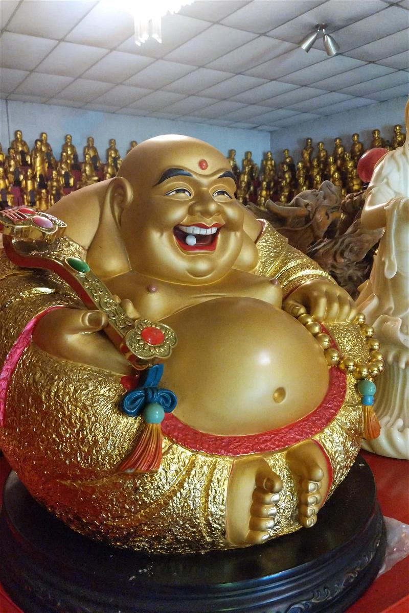A big fat golden smiling Buddha in a room of many tiny Buddhas along the walls at a temple