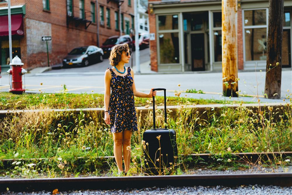 A woman standing next to a train track in New York City with a suitcase.