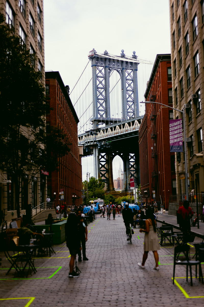 People are sitting on a sidewalk near a bridge in NYC.