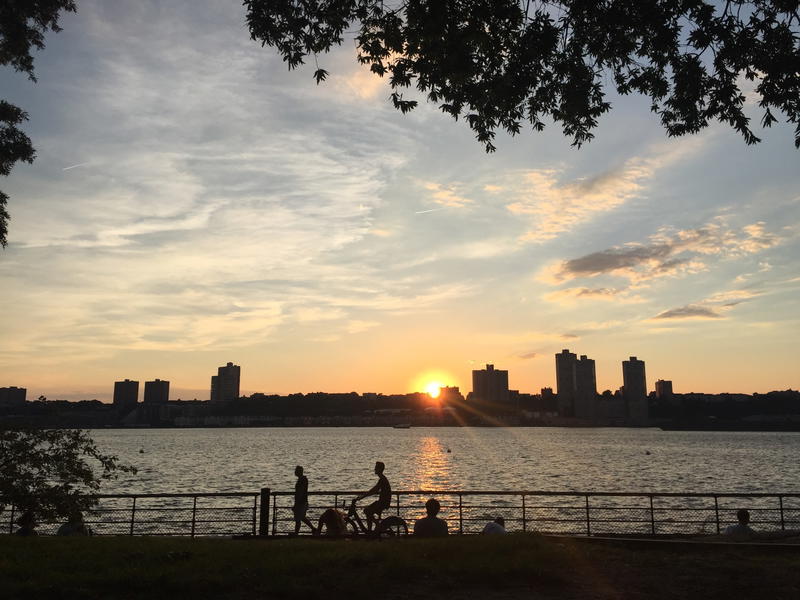 A group of people are sitting on a bench in NYC near a body of water as the sun is setting.